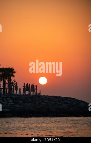 Un suggestivo e intenso tramonto arancione e rosso o alba sul mare, che delinea ombrelloni e palme su una costa rocciosa Foto Stock