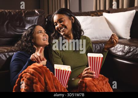 Assapora diverse amiche sedute sul pavimento accanto a un divano in pelle in coperta, con scatole di popcorn Foto Stock