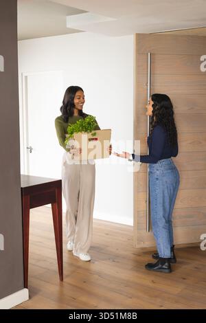 Diverse amiche si salutano a vicenda all'ingresso di casa, tenendo in mano una scatola di cartone con piante verdi Foto Stock