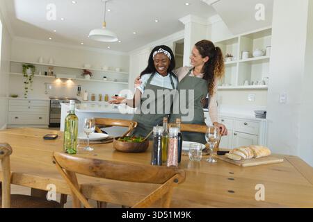Abbracciare diverse amiche in piedi a tavola nella cucina di casa, con insalata e bicchieri da vino Foto Stock