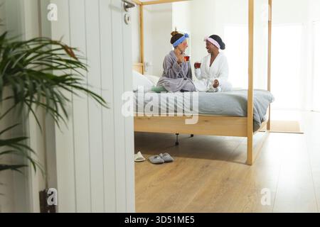 Chiacchierando con diverse amiche sedute su un letto a baldacchino nella moderna camera da letto, tenendo tazze di tè Foto Stock