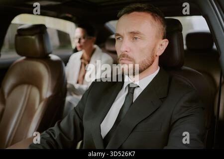 Uomo di mezza età alla guida di un'auto con donna d'affari seduta sul sedile posteriore Foto Stock