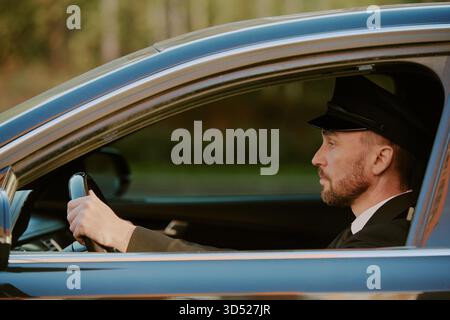Uomo di mezza età alla guida di un'auto indossando l'uniforme da autista Foto Stock