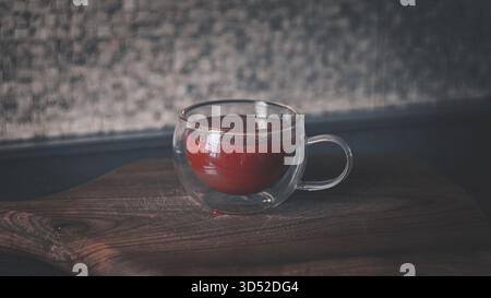 Una tazza di vetro trasparente piena di succo di pomodoro rosso brillante si trova su un piano di lavoro della cucina, illuminato da luce naturale che ne esalta la ricchezza Foto Stock