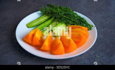 Verdure a fette, compresi cetrioli, pomodori arancioni e aneto, servite su un piatto su una superficie della cucina. La luce naturale enfatizza i colori vivaci e la Foto Stock