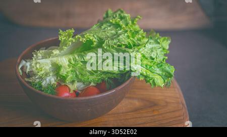 Una ciotola di foglie di insalata e ravanelli su una superficie rustica di legno. La luce solare calda filtra attraverso la finestra, proiettando ombre morbide e sottolineando la freschezza Foto Stock