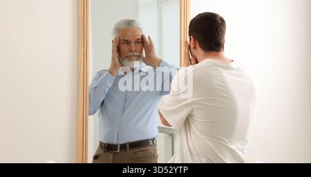Giovane uomo che si vede come anziano nello specchio. Concetto di personalità, passato e futuro, flusso del tempo, età Foto Stock