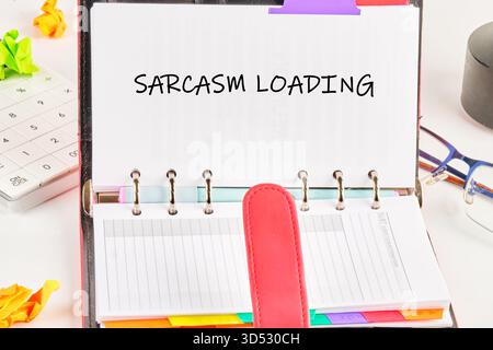 Un pianificatore giornaliero è aperto e mostra le parole caricamento sarcasmo. Accanto ad esso ci sono una calcolatrice, carta sgualcita e occhiali, che suggeriscono uno spazio di lavoro occupato e. Foto Stock