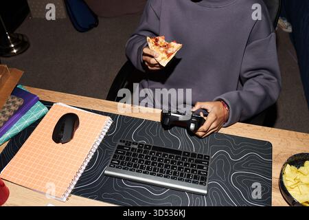 Adolescente seduto alla scrivania tenendo il controller di gioco in una mano e mangiando pizza con l'altra mano, tastiera e mouse visibili sulla scrivania, faccia parziale non visibile, probabilmente maschio, probabilmente caucasico Foto Stock