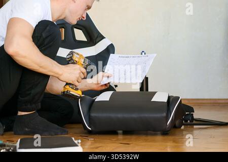 Uomo che utilizza un trapano elettrico su componenti per sedie da gioco con manuale nelle vicinanze. L'uomo inginocchiato aziona un trapano a batteria per fissare parti di un gioco in pelle nera Foto Stock