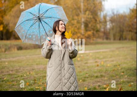 Donna felice con ombrello trasparente e foglia d'acero giallo che si gode una calda giornata autunnale nel parco Foto Stock