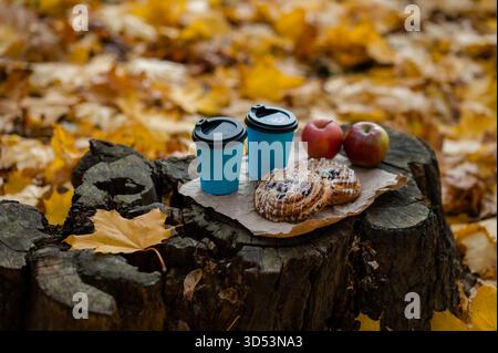 Scena autunnale concettuale che mostra due tazze, dolci e mele fresche disposte su un vecchio ceppo di alberi circondato da foglie autunnali cadute, evocando un ambiente accogliente Foto Stock