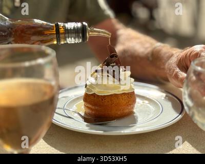 Primo piano di un uomo seduto all'aperto che versa rum su un dessert al rum baba (baba au rhum) Foto Stock