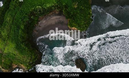La vista aerea delle verdeggianti scogliere incontra il turbolento mare turchese, dove le onde schiumose si infrangono contro la costa, Bali, Indonesia. Foto Stock