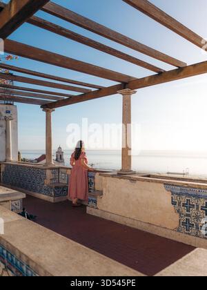 La vista di una donna guarda fuori da una terrazza piastrellata sotto un pergolato di legno verso il lontano mare e cielo blu, Lisbona, Lisbona, Portogallo. Foto Stock