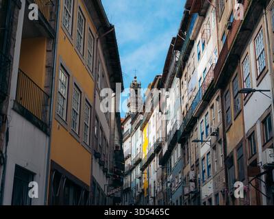 Veduta della stretta strada incorniciata da alti edifici con facciate colorate che conducono a una torre della chiesa contro un cielo blu, Porto, quartiere di Porto, Portogallo. Foto Stock