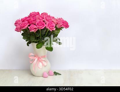 Mazzo di fiori rosa rose in una vaso di porcellana bianca e cuori rosa su un tavolo bianco su sfondo chiaro con spazio per il testo Foto Stock