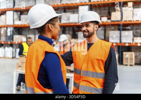 Due diversi addetti al magazzino che indossano elmetti e giubbotti di sicurezza sorridono con fiducia e collaborano in un centro di distribuzione logistica, demoniaco Foto Stock