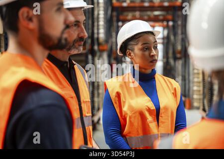 Professionisti del settore che indossano giubbotti di sicurezza e elmetti discutono di piani di lavoro e operazioni in un moderno magazzino logistico, che rappresenta il lavoro di squadra Foto Stock