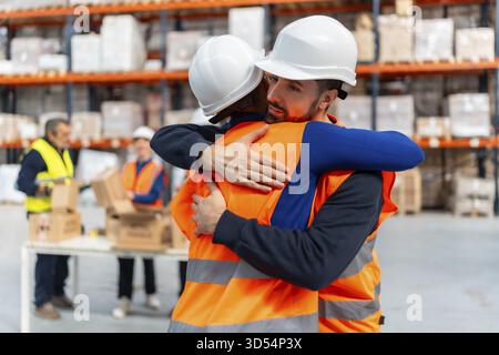 Due diversi operatori di magazzino che indossano giubbotti di sicurezza e cappelli rigidi che abbracciano in una struttura logistica, simboleggiano l'unità, il supporto e un forte senso di c Foto Stock