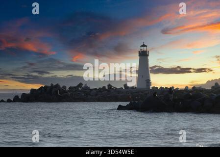 Un faro si erge graziosamente su una scogliera rocciosa, sagomato contro un vibrante cielo al tramonto pieno di sfumature di arancione, rosa e viola. Calma Foto Stock