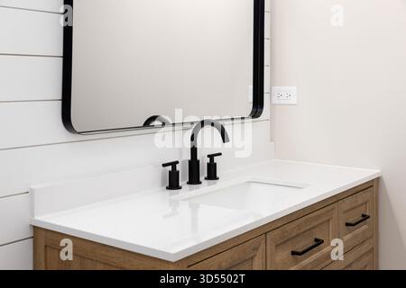 Un bagno con armadi in quercia bianca, rubinetto nero su un ripiano in marmo bianco e uno specchio nero su una parete in shiplap bianco. Foto Stock