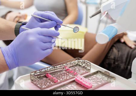 Primo piano di un medico con i guanti che tiene uno specchio e un esploratore sopra una cassetta di sterilizzazione con un paziente sulla sedia, mostrando il flusso di lavoro degli esami dentistici Foto Stock