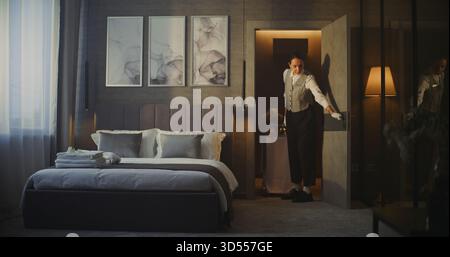 Il giovane dipendente dell'hotel in uniforme porta il carrello del servizio in camera in una camera moderna in un hotel di lusso. La donna offre squisita colazione e fiori freschi in camera. Comfort e ospitalità di qualità. Foto Stock
