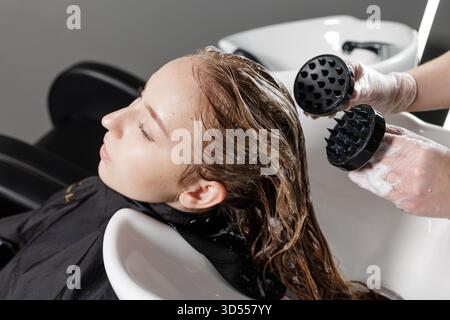 Lo stylist con guanti utilizza un massaggiatore per cuoio capelluto in silicone e una spazzola per shampoo durante il lavaggio del salone, migliorando la pulizia, la circolazione e la cura rilassante del centro benessere della testina. Foto Stock