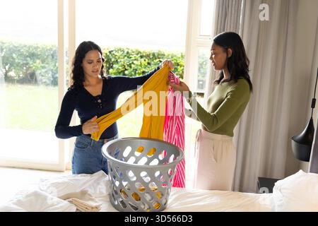 Lavorate diverse amiche con lavanderia pieghevole sul letto a casa, con cestino grigio e abito a righe Foto Stock