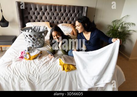 Diverse amiche, lavanderia pieghevole sul letto della camera da letto, con smartphone, cestino grigio Foto Stock