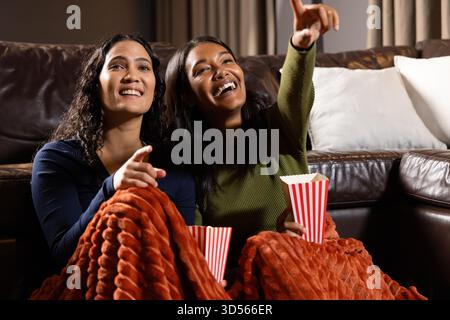 Indicando diverse amiche che siedono nel soggiorno con una coperta arancione, tenendo in mano vasche di popcorn Foto Stock