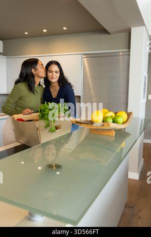 Diverse partner femminili si baciano mentre si appoggiano su un'isola di vetro nella cucina di casa, con casse di prodotti Foto Stock