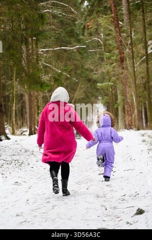 Mamma e figlia corrono con gioia lungo il sentiero innevato della foresta, vista dal retro. Avventure invernali in famiglia nella foresta invernale. Concetti di camminata invernale, C. Foto Stock