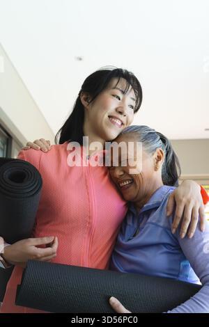 Abbracciare madre e figlia cinesi tenendo in mano tappetini neri da yoga in palestra, preparandosi per la lezione Foto Stock
