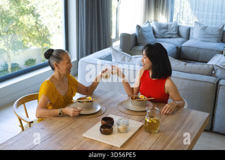 Madre e figlia asiatica cinesi sorridenti mangiano insalata di noodle nella moderna sala da pranzo, con tè freddo Foto Stock