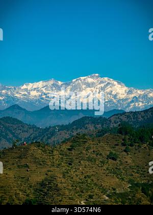 Splendida vista delle montagne himalayane con cime innevate sotto un cielo azzurro. Verdi colline e piccole case di villaggio nella valle creano un paesaggio naturale tranquillo e panoramico. Foto Stock