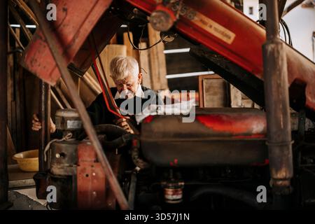 Uomo anziano che ripara un trattore d'epoca in un'officina rustica Foto Stock