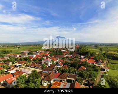 Vista aerea di un vivace villaggio annidato tra campi di riso color smeraldo, dominato dal maestoso Monte Merapi sotto un cielo azzurro, Klaten Regency, Ja Foto Stock