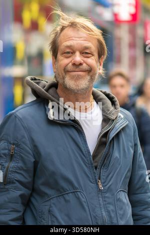 Un uomo di 50-55enne con una giacca sorride mentre si trova su una strada trafficata della città tra i passanti Foto Stock