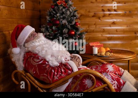 Babbo Natale dorme tranquillamente su una sedia, con un albero di Natale festoso Foto Stock