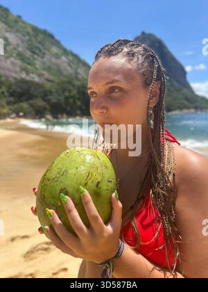 Rio de Janeiro . Brasile. Giovane donna in bikini rosso con capelli intrecciati che beve da una noce di cocco fresca su una spiaggia tropicale soleggiata, rilassata e allegra. Foto Stock