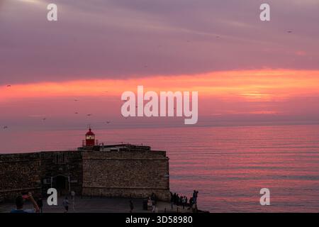 Un vivido tramonto bagna il cielo e l'oceano in calde tonalità rosa e arancio dietro lo storico forte de Sao Miguel Arcanjo a Nazare, Portogallo Foto Stock