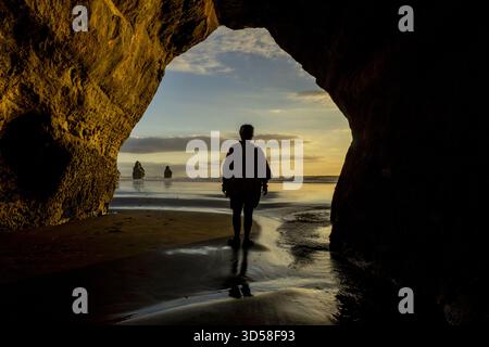 Vista di una figura sagomata che cammina sulla spiaggia attraverso un arco naturale verso formazioni rocciose lontane sotto un cielo morbido e dorato, Tongaporutu e Tar Foto Stock