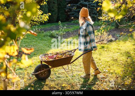 Giardiniere che spinge carriola con rastrello e foglie cadute per il compost. Giardinaggio autunnale. La donna sta rastrellando le foglie in giardino Foto Stock