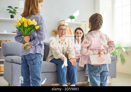 Due bambini nascondono tulipani e carta dietro le spalle mentre si congratulano con la madre felice e la nonna. Foto Stock