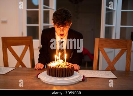 Un ragazzo di 12 anni fa fa saltare le candele sulla torta di compleanno indossando l'uniforme scolastica, dopo essere tornato a casa da scuola, Inghilterra, Regno Unito Foto Stock