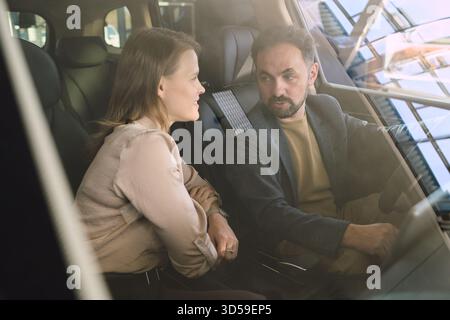 Giovane donna adulta caucasica e giovane adulto mediorientale seduto all'interno dell'auto a discutere le caratteristiche del veicolo presso la concessionaria, entrambi affrontati con espressioni attente Foto Stock