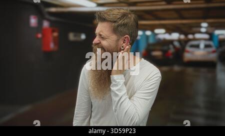 L'uomo con barba folta e camicia bianca informale tiene mano a mano nell'edificio pieno di auto sfocate che riflettono le luci sopra la testa; disagio. Foto Stock