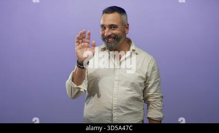 Uomo ispanico di mezza età con barba al sale e al pepe sorridente in uno studio viola con camicia in lino beige e maniche arrotolate; amichevole fiducia. Foto Stock